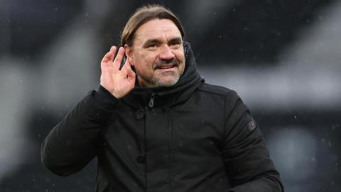 Leeds United manager Daniel Farke, dressed in a black winter coat and cupping his hand to his ear