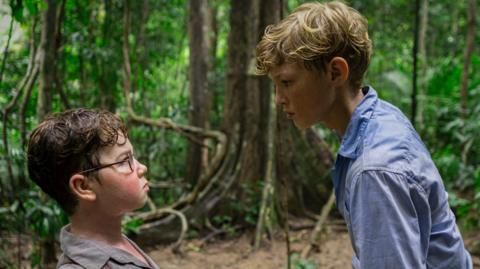 A young boy with brown hair and glasses looks up at a blonde haired boy in a blue shirt. They are in a wooded area of a tropical island.