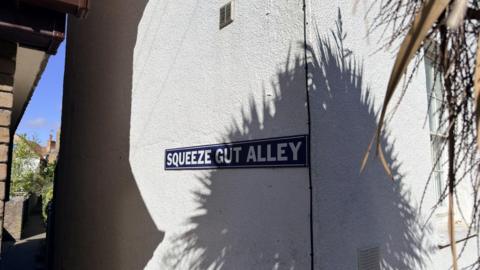 A black and white sign, reading Squeeze Gut Alley, besides a passageway. 