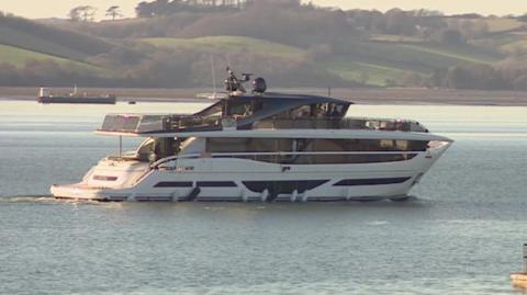 A blue and white yacht on the water with rolling green hills in the background.