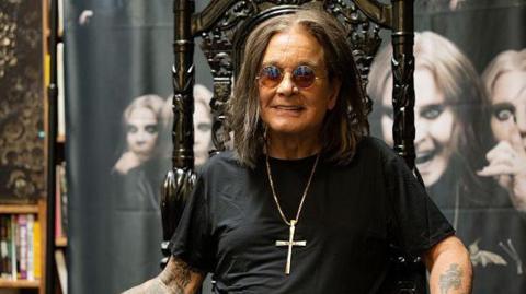 Ozzy Osbourne, who has shoulder-length brown hair, is wearing tinted round glasses, a black T-shirt and a large silver cross on a chain around his neck. He has tattoos on his arms and is sitting in an ornate dark chair in front of a screen with black and white images of his face.