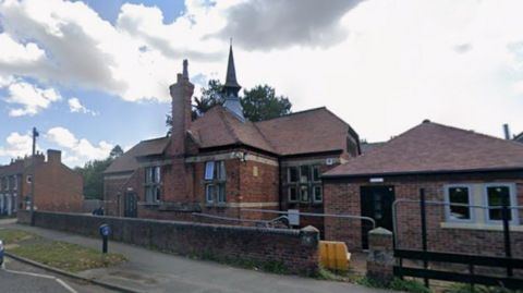 A red brick building behind a low wall with a tall chimney and a pointed tower on the roof