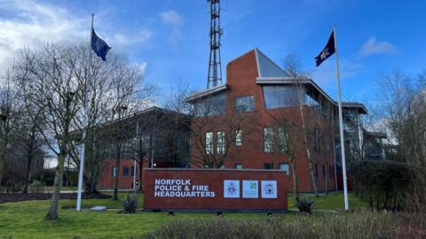 Exterior of police headquarters in Wymondham. The building is made mostly of red brick with a lawned frontage and birch trees. Two flags on masts are erected on the lawn.