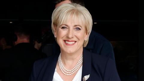 Heather Humphreys is wearing a white top and navy blazer. She is wearing 3 pearl chains around her nice. She has short blonde hair. Behind her are other people but it is blurry. 