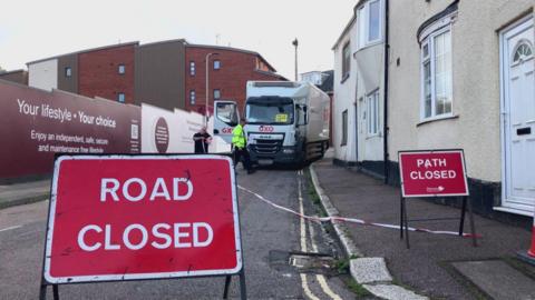 Lorry stuck on narrow road after scaffolding collision. A red road closed sign is blocking the road. A red path closed sign has shut the pavement to pedestrians. Residential buildings line the street. Two men are at the scene.