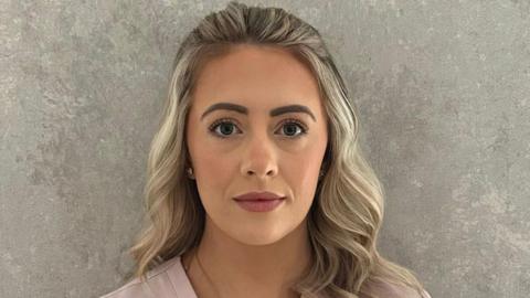 Claire Harkin has long wavy blonde hair. She has some mascara and pink lipstick on and is wearing a pink top.