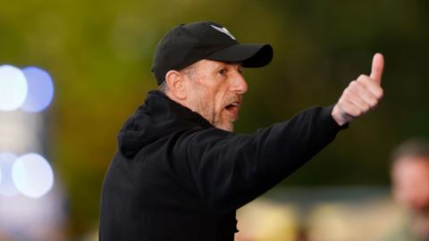 gary rowett puts his right arm out to the side with his thumb up and has his mouth open speaking. he wears a black hoodie and black cap