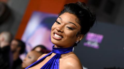 Megan Thee Stallion pictured at the premiere of her documentary in October. Megan wears a royal blue halterneck dress and smiles at the camera. Her hair is styled in a bun and guests can be seen in the background. 