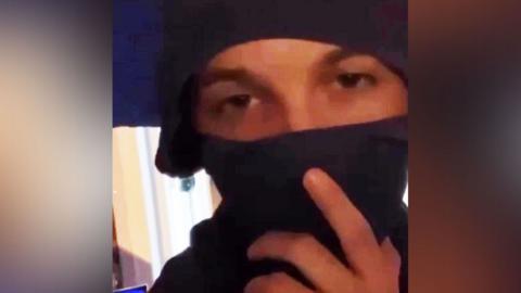 A young man looking into the camera, most of his face is obscured by a piece of clothing which he wears like a mask. He holds the garment over his mouth.