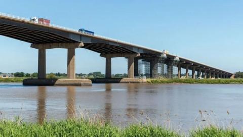 View from the riverbank of the piers of the Ouse Bridge reflecting in the water, with three lorries making their way over the bridge