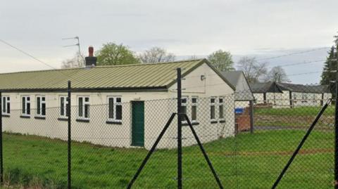 A number of long one-storey white buildings with sloped green rooves on a grassy area with black metal fencing surrounding them