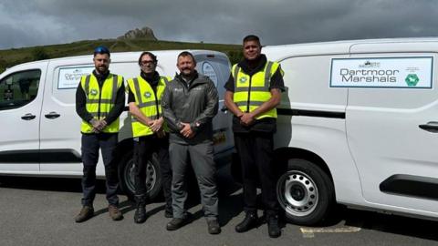 Three Dartmoor Marshals wearing yellow high-vis jackets stand with Dartmoor's head ranger Simon Lee in front of a tor on the moor and two vans with Dartmoor Marshals logos on them. It is an overcast day with grey clouds looming in the sky.