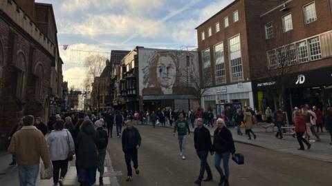 A general view of a busy street in Exeter. Crowds of people are walking along the pavements. Some people are crossing the road. Tall buildings line either side of the street. A mural of a woman's face is painted on the side of one building. 