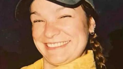 Clare Dupree, a woman with a big smile wearing a baseball cap and yellow coat. Her hair is braided into a plait and falling behind her shoulder.