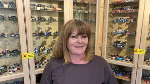A woman standing indoors in front of wooden display cabinets with glass shelves. The cabinets are filled with numerous small model racing cars arranged neatly in rows. Yellow signs reading “PLEASE DO NOT TOUCH” are visible on the display cases, indicating a collection or exhibition setting.
