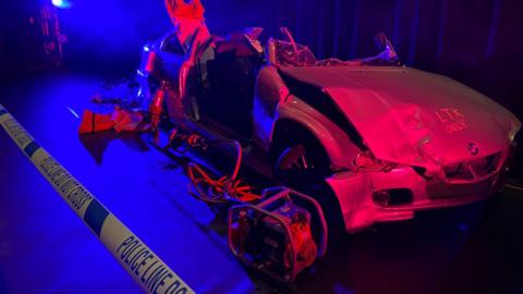 A crashed car behind police line "do not cross" tape. The car is part of the presentation exhibition. It is mangled and with red light being shone on it. The roof is missing and one of the doors has been taken off 