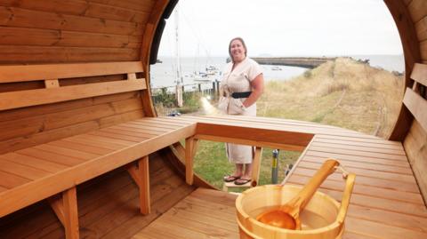 The image shows the inside of a wooden style sauna, Kathryn Donavan, the owner of the sauna is stood just outside. She's wearing a beige dress with a black belt and a pair of black flip flops. She has short blonde hair.