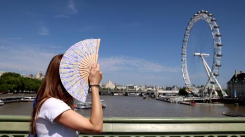 A woman with her face covered by her hand-held fan, with the River Thames and London Eye in the background