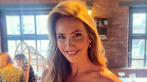 Claire Laybourne sits in a restaurant with a brick wall. She has long, blonde highlighted and brown hair and wears gold earrings. There is a section of exposed brick wall between to large windows behind her and, slightly out of focus, two women are seated at another table.
