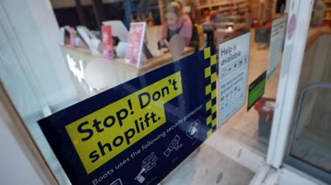 A sign in a shop window says 'Stop! Don't shoplift. Boots uses the following security protection'. A blurred out customer can be seen using a self service till inside the store.