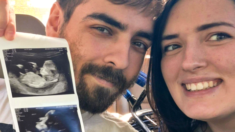 A man with short dark brown hair and a beard sits next to a woman with shoulder length brown hair on a hospital bed. He is holding up baby scan images to show the camera.