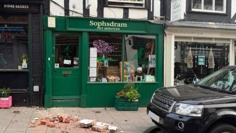 Rubble lies on the pavement outside a green painted shop frontage with Sophsdram Pet Services written above the window. A black Land Rover can be seen parked alongside it, and other ships either side, including a beauty salon.
