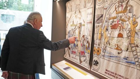 King Charles pointing at a colourful tapestry depicting "Bruce's siege of Carlisle and beginning of little ice age".