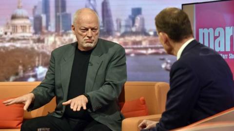 David Gilmour sitting on orange sofa holding out his hands while being interviewed by the tv's Andrew Marr, who has his back to the camera