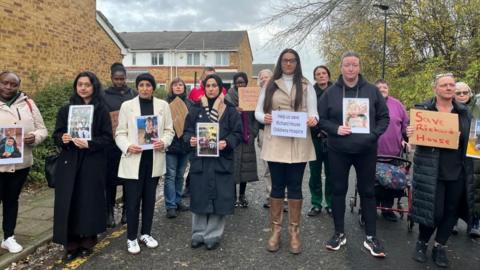 A number of parents and carers stand in the road holding signs and pictures of children who have been supported by Richard House.  Some signs read "Save Richard House"