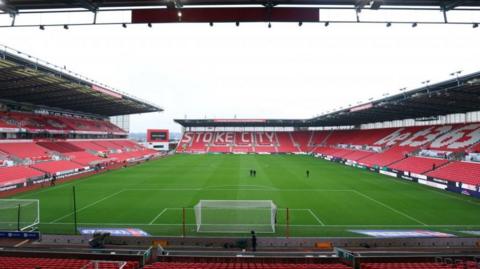 The view onto the pitch from the stands of a large football ground. The seats are red, with Stoke City picked out in white on the opposite side stand