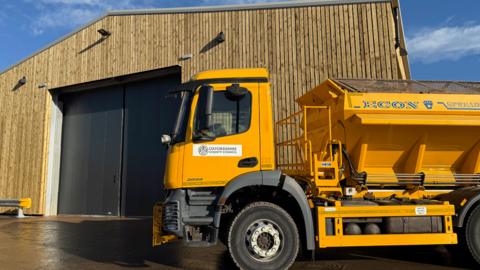 A gritting truck is parked outside the new salt barn. The truck is yellow, with a large container on its back for carrying salt. The large barn looks wooden in appearance, with great big metal doors, large enough for vehicles to drive through.