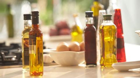A collection of bottles of cooking oils and vinegars on a kitchen counter. There is a bowl of eggs in the centre and a gas hob in the left background.