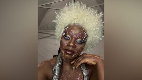 A person in bold avant‑garde makeup with bright blue eyes, a large white beaded wig, and metallic costume accessories, standing in a tent-like backstage setting