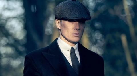 The actor Cillian Murphy dressed as Tommy Shelby, the character he plays in the drama Peaky Blinders. He is wearing a black and white tweed flat cap, black jacket, white shirt, and a tie. There are trees in the background.