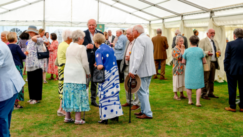 A wide shot of a garden party. There are hundreds of people gathered inside a large marquee.