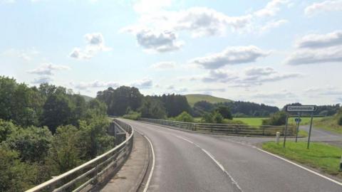 A road with barriers on either side. There are also trees in full leaf on both sides of the road, and a hill in the distance. There is a road sign on the right pointing to Commonside.