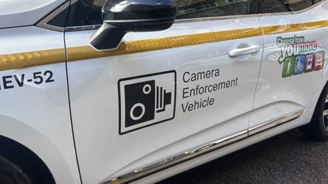 Branding and markings on the side of a white car, including a speed camera sign and the words Camera Enforcement Vehicle