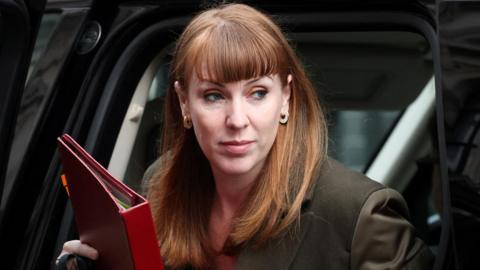 Angela Rayner, a woman with short red hair and a fringe wearing a brown jacket, steps out of a black car holding a red folder.