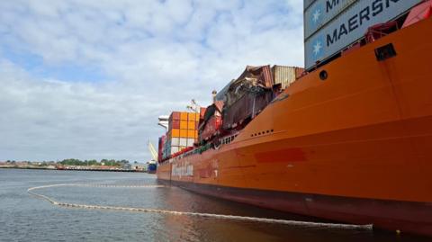 A large orange container ship with a white boom - a barrier - in a semi-circle in the water next to it. On the ship are several crushed containers. Stacks of undamaged shipping containers can be seen on the of the ship. 