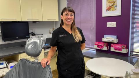 A woman in her 30s in black scrubs branded with Chertsey house on the left hand side. She is long brunette hair that gets blonder towards the bottom. She is smiling as she leans on a dentist chair in the surgery.