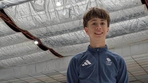 Edward Solovyov stand on the ice at Whitley Bay Ice Rink, he is wearing his new dusty-blue Team GB official teamwear. Behind him, silver foil insulation hangs from the roof.