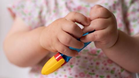 A baby's hands pictured holding a chunky pen that is yellow, red and blue. They are wearing a pink and green floral top. 