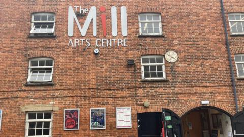 The front facade of the mill. It is red brick and has lots of windows. There are posters advertising upcoming shows and a big sign saying 'The Mill Arts Centre'.