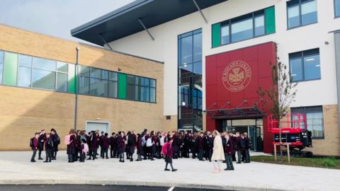 Outside St Ronan's College. The main building is white with large windows and a red section for the school sign. Another building to she side has beige bricks and large windows. Pupils in burgundy uniforms are gathering outside the entrance.