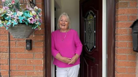 Pauline Manning is standing at the front door of her home.  She has a faux flower hanging basket to left along with a ring doorbell. She is wearing a bright pink top, white trousers and is smiling at the camera. 