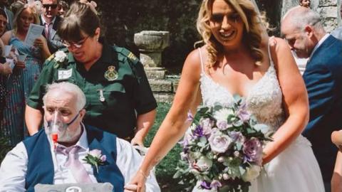 Wayne is in a wheelchair, wearing an oxygen mask. He is being pushed down the "aisle" of the outdoor wedding by a paramedic in uniform. His daughter is dressed in a wedding dress with bouquet and is holding his hand.