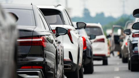 The image shows cars lined up on a road in a traffic jam. In the distance, an open car door can be seen on the left hand side.