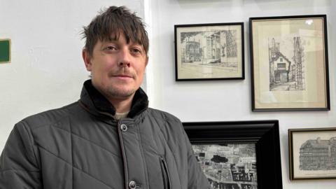 Scott Duffin is wearing a dark grey coat, with collar turned up. He is standing in front of prints which ar part of the Coventry Market exhibition