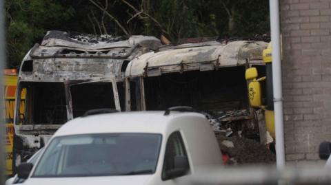 The picture shows two burn-out lorries at an industrial estate, with a white van in the foreground