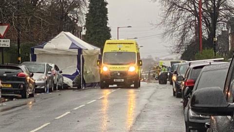 A rainsoaked road, there are park cars either side and a yellow ambulance with its lights on. It is next to a blue and white tent.